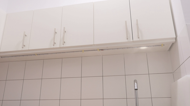 The camera moving from left to right and shooting a white cupboard under the ceiling. White luxury tiles are on the walls under the cupboards. The camera focuses on a sink with metallic faucet and empty tabletop. - Footage, Video