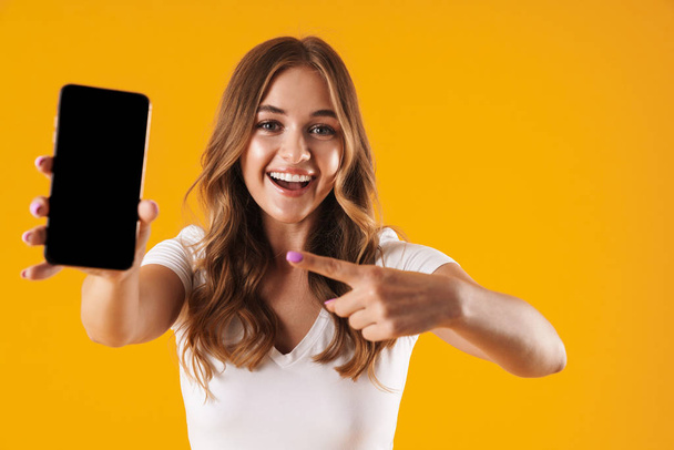 Image closeup of young caucasian woman wearing basic t-shirt rejoicing while pointing finger at cellphone - Foto, immagini