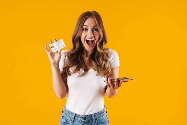 Photo of pretty young woman rejoicing while holding credit card and smartphone isolated over yellow background - Fotografie, Obrázek