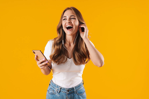 Image closeup of happy caucasian woman wearing basic t-shirt smiling while listening to music with cellphone and earphones - Fotó, kép