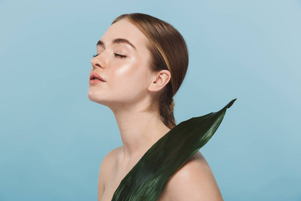 Beautiful young woman take care of her skin isolated over blue wall background posing with leaf flower. - Foto, Imagen