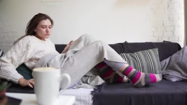 A young woman in warm multi-colored socks and pajamas sits more comfortably on a cozy sofa. Hygge concept - Filmati, video