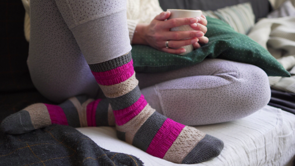 Růžové a hnědé vlažné ponožky zblízka-žena sedí s bílým šálkem horkého nápoje na pohovce s kostkami a polštáři. Pojem Lagom nebo hygge-komfort a jednoduchost - Záběry, video