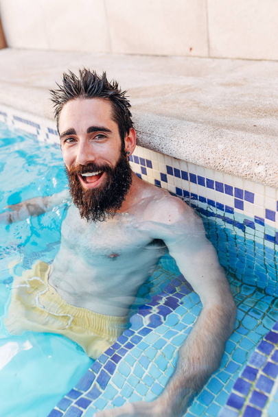 Portrait of a young man with a beard smiling and sitting in the poolside. Holiday concept - Fotó, kép