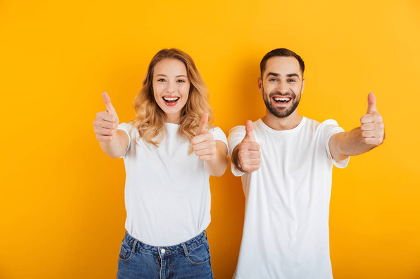 Portrait of cheerful young couple man and woman in basic t-shirts smiling and showing thumbs up at camera - Φωτογραφία, εικόνα