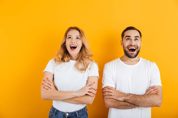 Portrait of excited young couple man and woman in basic t-shirts wondering and looking upward with arms crossed - Фото, зображення