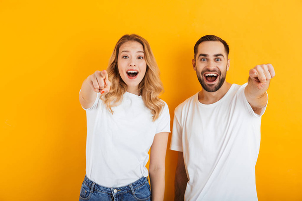 Portrait of happy young couple man and woman in basic t-shirts smiling and pointing fingers at camera - Foto, immagini
