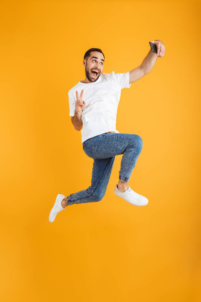 Full length photo of happy caucasian man having beard taking selfie on cellphone and showing peace sign - Foto, afbeelding