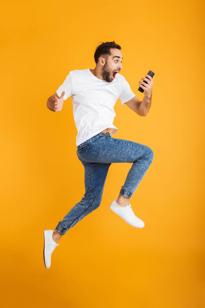 Full length photo of shocked caucasian man having beard wondering and holding cellphone while jumping - Fotografie, Obrázek