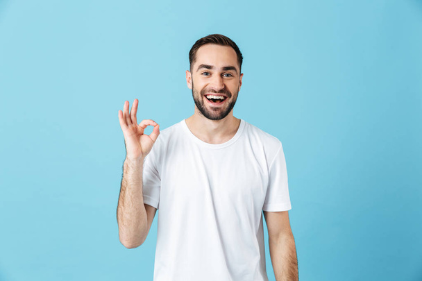 Smiling young happy bearded man posing isolated over blue wall background showing okay gesture. Summer concept. - Fotografie, Obrázek