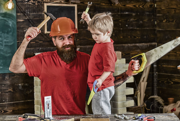 Αγόρι, παιδί χαρούμενο κατέχει παιχνίδι saw, έχει τη διασκέδαση ενώ handcrafting με τον μπαμπά. Πατέρα, του γονέα με γενειάδα στο προστατευτικό κράνος διδασκαλία γιο να χρησιμοποιούν διαφορετικά εργαλεία στο σχολικό εργαστήριο. Πατρότητα έννοια - Φωτογραφία, εικόνα
