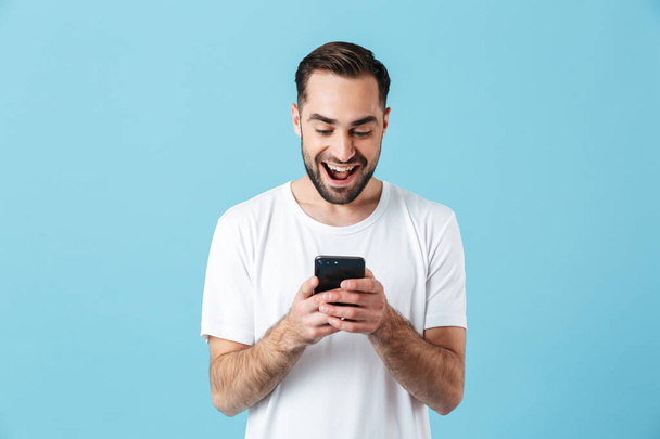 aufgeregte junge glückliche bärtige Mann posiert isoliert über blaue Wand Hintergrund mit Handy-Chat. - Foto, Bild