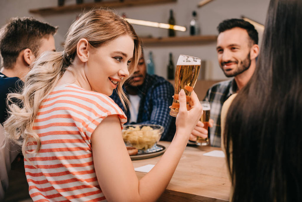 selektywne skupienie uśmiechniętej młodej kobiety trzymając szklankę piwa siedząc z wielokulturowym znajomym w pubu - Zdjęcie, obraz