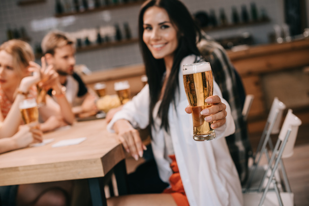 selectieve focus van vrolijke jonge vrouw die glas licht bier vasthoudt en naar de camera kijkt terwijl je in de buurt van vrienden zit in Bar - Foto, afbeelding