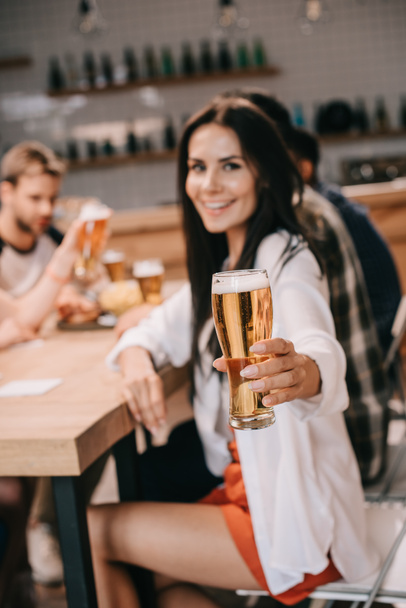 enfoque selectivo de la joven mujer sosteniendo un vaso de cerveza y sonriendo a la cámara mientras se sienta en el pub junto con sus amigos
 - Foto, Imagen