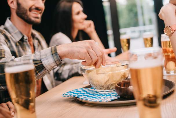 partial view of man taking chips from bowl while sitting with friends in pub - Foto, Imagen