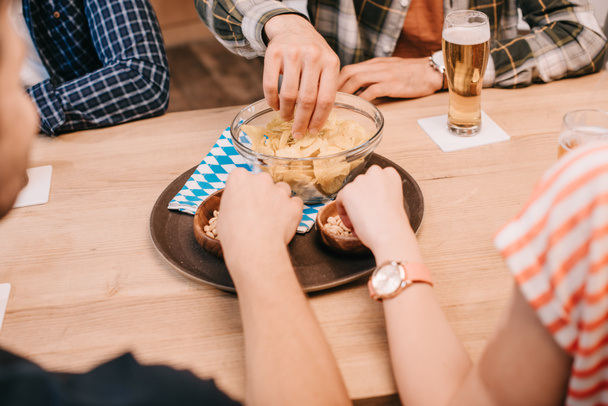 partial view of friends taking snacks from bowls while sitting together in pub - Photo, Image