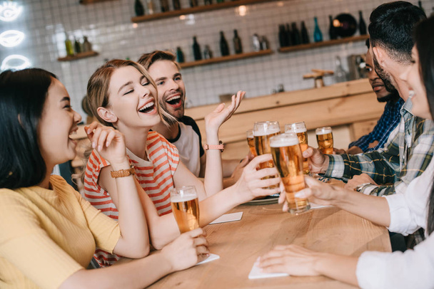 cheerful multicultural friends talking and gesturing while drinking beer in pub together - Фото, зображення