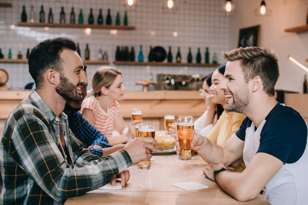 alegres amigos multiculturales hablando y bebiendo cerveza ligera en el pub
 - Foto, Imagen