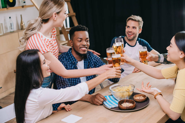 cheerful multicultural friends clinking glasses of light beer in pub - Photo, Image