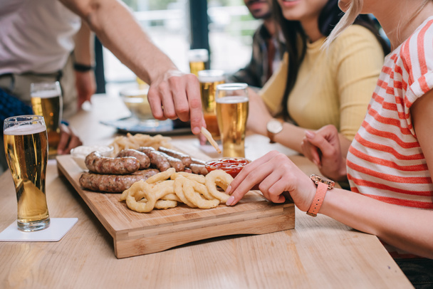 vista parcial de amigos multiculturales sentados en el pub cerca de la bandeja con aros de cebolla frita, salchichas y papas fritas
 - Foto, Imagen