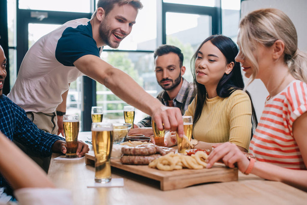vrolijke multiculturele vrienden zittend in de pub in de buurt van tray met gebakken uienringen, worstjes en frietjes - Foto, afbeelding