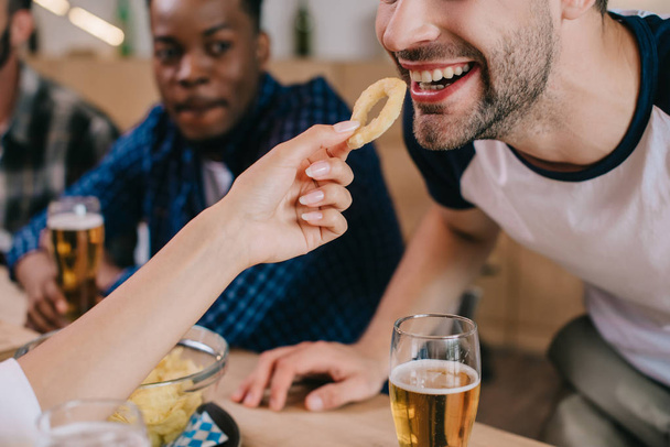 vista recortada de la mujer alimentando hombre alegre con anillo de cebolla frita mientras está sentado en el pub con amigos
 - Foto, Imagen