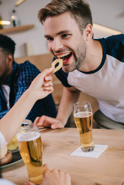 vista parcial de la mujer alimentando hombre alegre con anillo de cebolla frita mientras está sentado en el pub con amigos multiculturales
 - Foto, Imagen