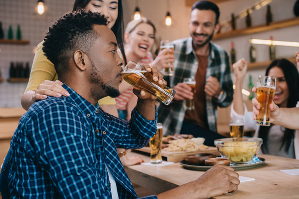 enfoque selectivo del joven afroamericano bebiendo cerveza ligera mientras pasa tiempo en el pub junto con amigos alegres multiculturales
 - Foto, Imagen