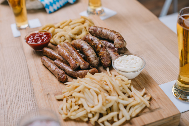 ξύλινο δίσκο με τηγανητά λουκάνικα, κρεμμύδια, τηγανητές πατάτες και σάλτσες κοντά σε ποτήρια με ελαφριά μπίρα στην παμπ - Φωτογραφία, εικόνα