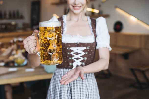 cropped view of  waitress in german national costume holding mug of light beer  - Фото, изображение
