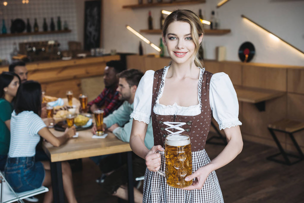 gyönyörű pincérnő a német nemzeti ruha gazdaság bögre könnyű sör és mosolygott a kamera - Fotó, kép