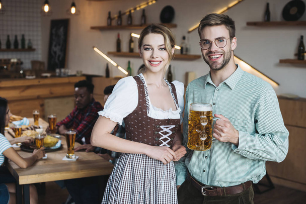 bella cameriera in costume nazionale tedesco in piedi vicino al giovane uomo che tiene una tazza di birra leggera e sorride alla macchina fotografica
 - Foto, immagini