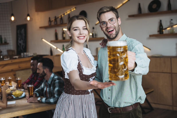 selektivní zaměření pohledného mladého muže, který drží džbánek lehkého piva, zatímco stojí u krásné servírce v tradičním německém kostýmu - Fotografie, Obrázek