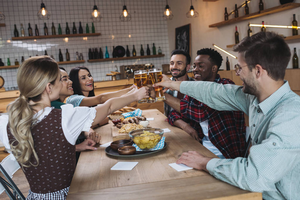 cheerful multicultural friends clinking glasses of light beer while celebrating octoberfest in pub - 写真・画像