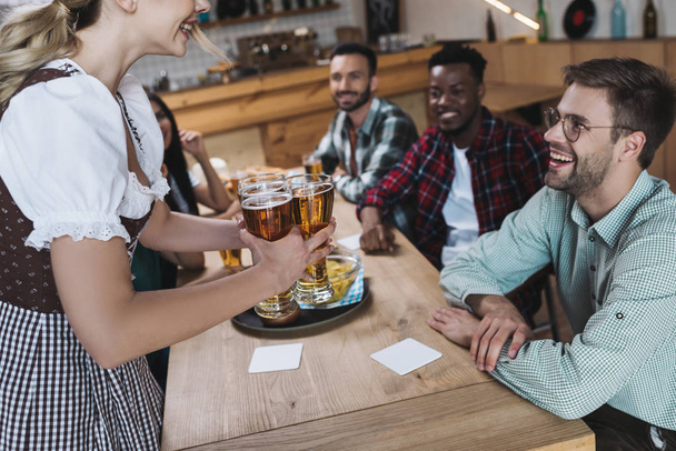 abgeschnittene Ansicht einer Kellnerin mit Gläsern leichten Bieres in der Nähe multikultureller Freunde - Foto, Bild