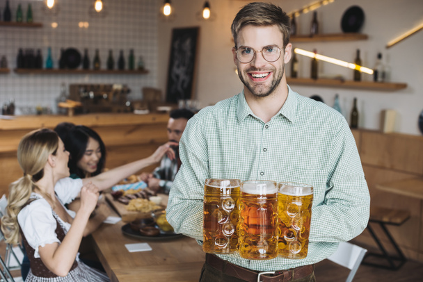 Красивый молодой человек держит кружки с пивом и улыбается в камеру
 - Фото, изображение