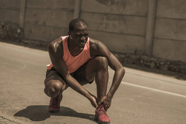 young athletic and fit black African American runner in pain holding his ankle after suffering medical problem with injuried tendon or ligament during urban running workout - 写真・画像