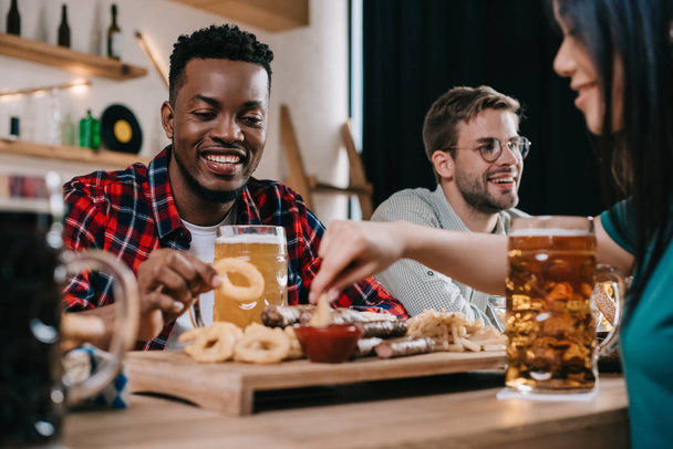 smiling multicultural friends eating fried onion rings with ketchup in pub - Photo, Image