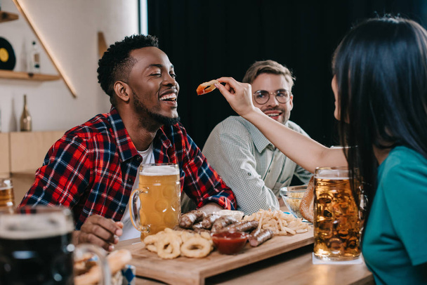 back view of young woman feeding african american man with fried onion ring while celebrating octoberfest in pub - Foto, Imagem