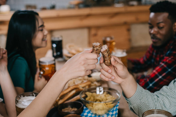 vista recortada de la joven mujer y la mujer sosteniendo salchicha frita mientras celebra octoberfest con amigos multiculturales
 - Foto, Imagen
