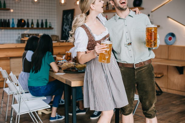 vista ritagliata di uomo e donna allegro in costumi tradizionali tedeschi che tengono tazze di birra
  - Foto, immagini