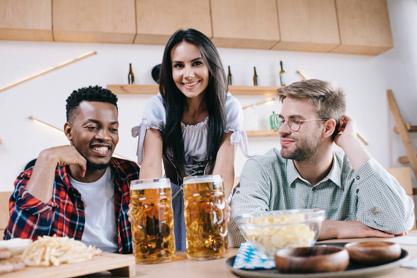 aantrekkelijke serveerster in traditioneel Duits kostuum serveren bier voor multiculturele vrienden in pub - Foto, afbeelding