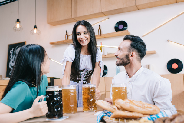 gut gelaunte Kellnerin in Tracht serviert Bier für Mann und Frau in der Kneipe - Foto, Bild