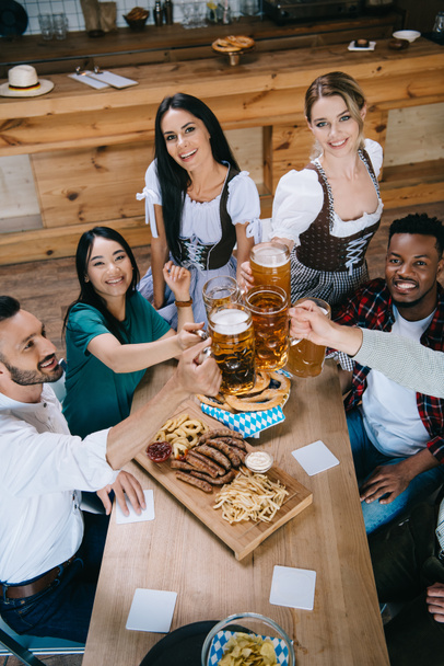 hermosas camareras en trajes tradicionales alemanes tazas tintineo de cerveza con amigos multiculturales
 - Foto, Imagen