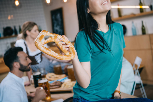 cropped view of smiling asian girl holding pretzel while celebrating octoberfest with multicultural friends - Photo, Image