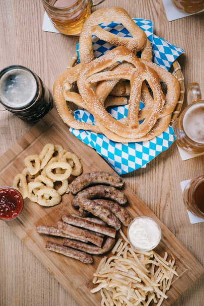 top view of fried sausages, onion rings, french fries, pretzels and mugs with beer on wooden table in pub - Photo, Image