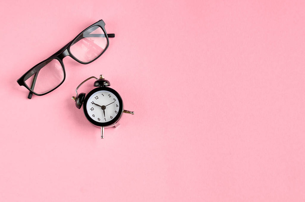 Black glasses and alarm clock on pink background composition. - Photo, Image