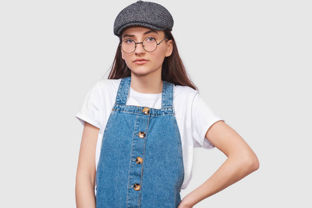 Studio image of serious young woman wears transparent trendy eyewear, casual white t-shirt, denim dress, trendy gray cap, looks seriously directly into camera, poses on white wall. People emotions - Фото, зображення