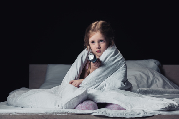 frightened child sitting under blanket and holding flashlight isolated on black - Foto, Imagem
